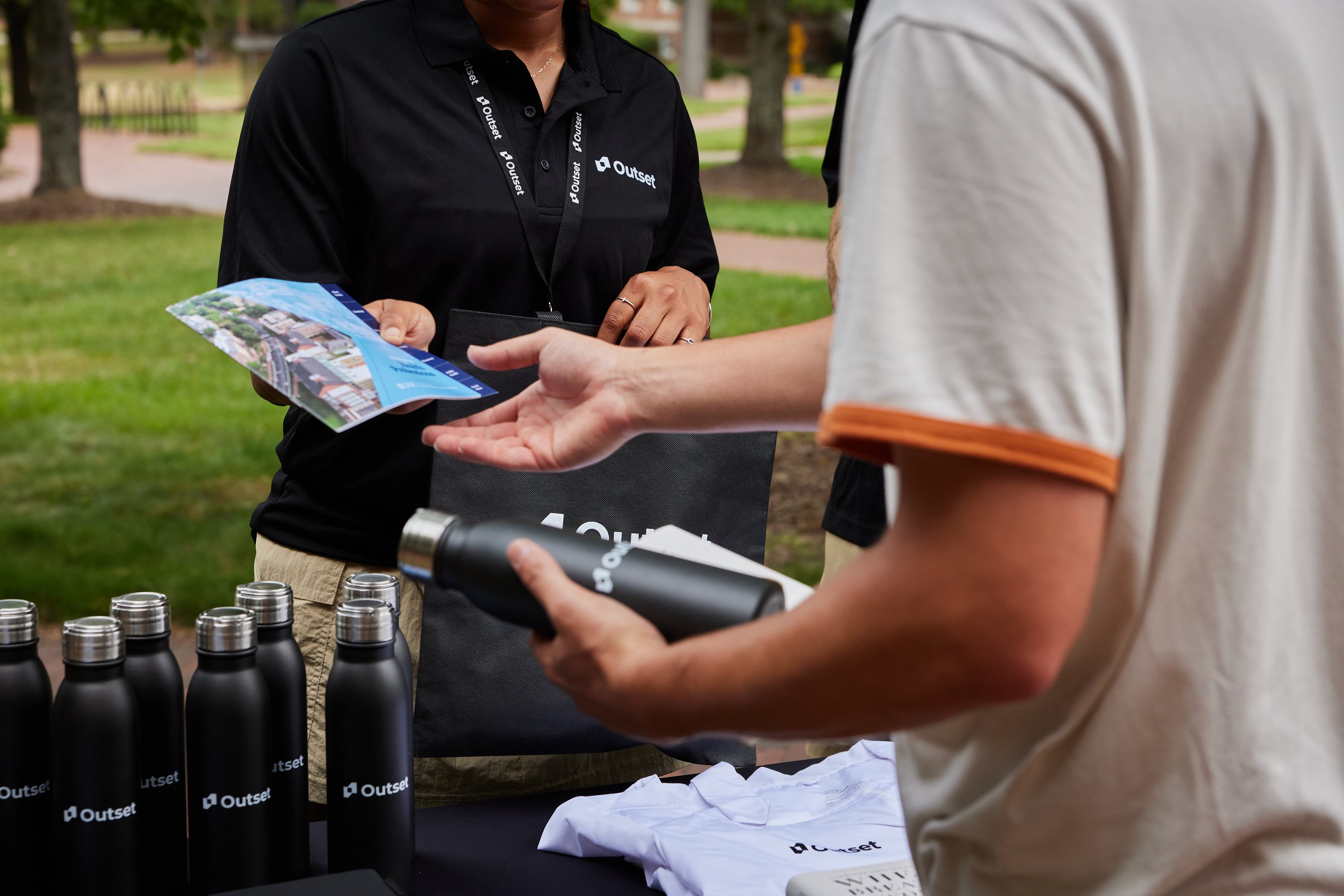 Outset staff member hands brochure to a student holding a black "Outset" water bottle at an outdoor table with more bottles and shirts, surrounded by a grassy park setting.