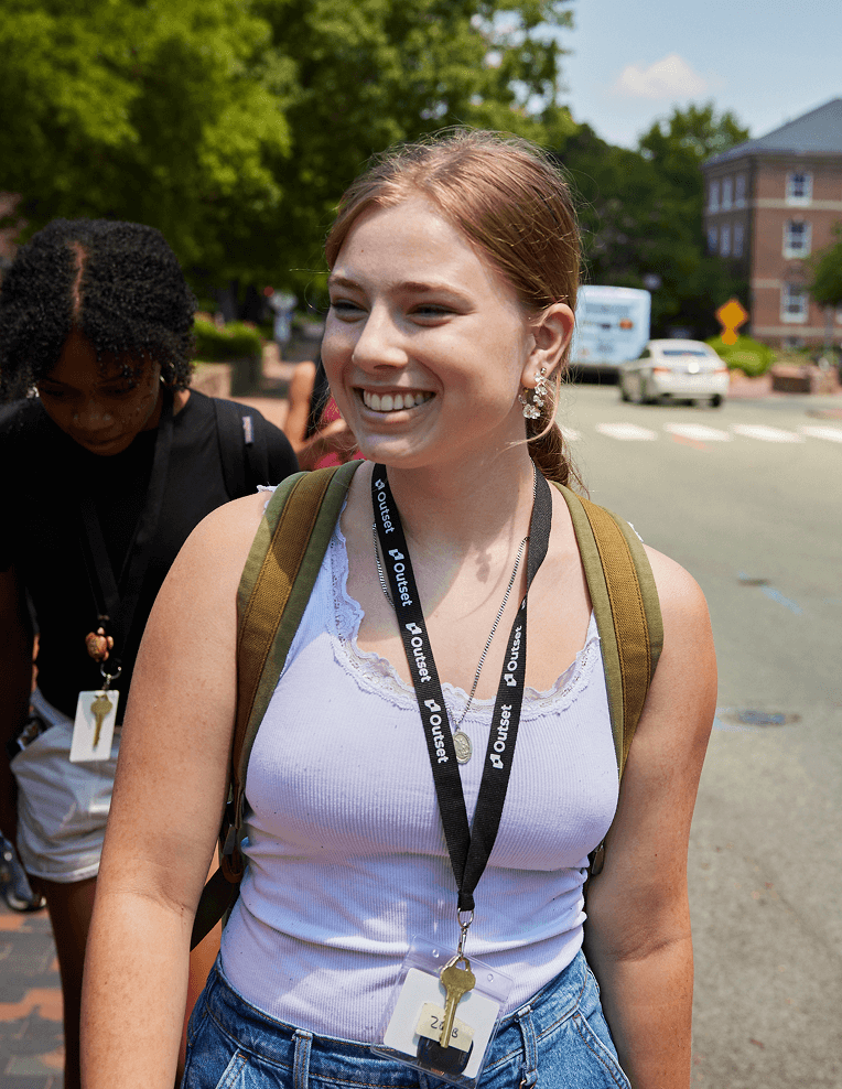 A future medical student wearing a backpack and a lanyard with a badge smiles while walking outdoors on a sunny street, surrounded by trees and buildings.