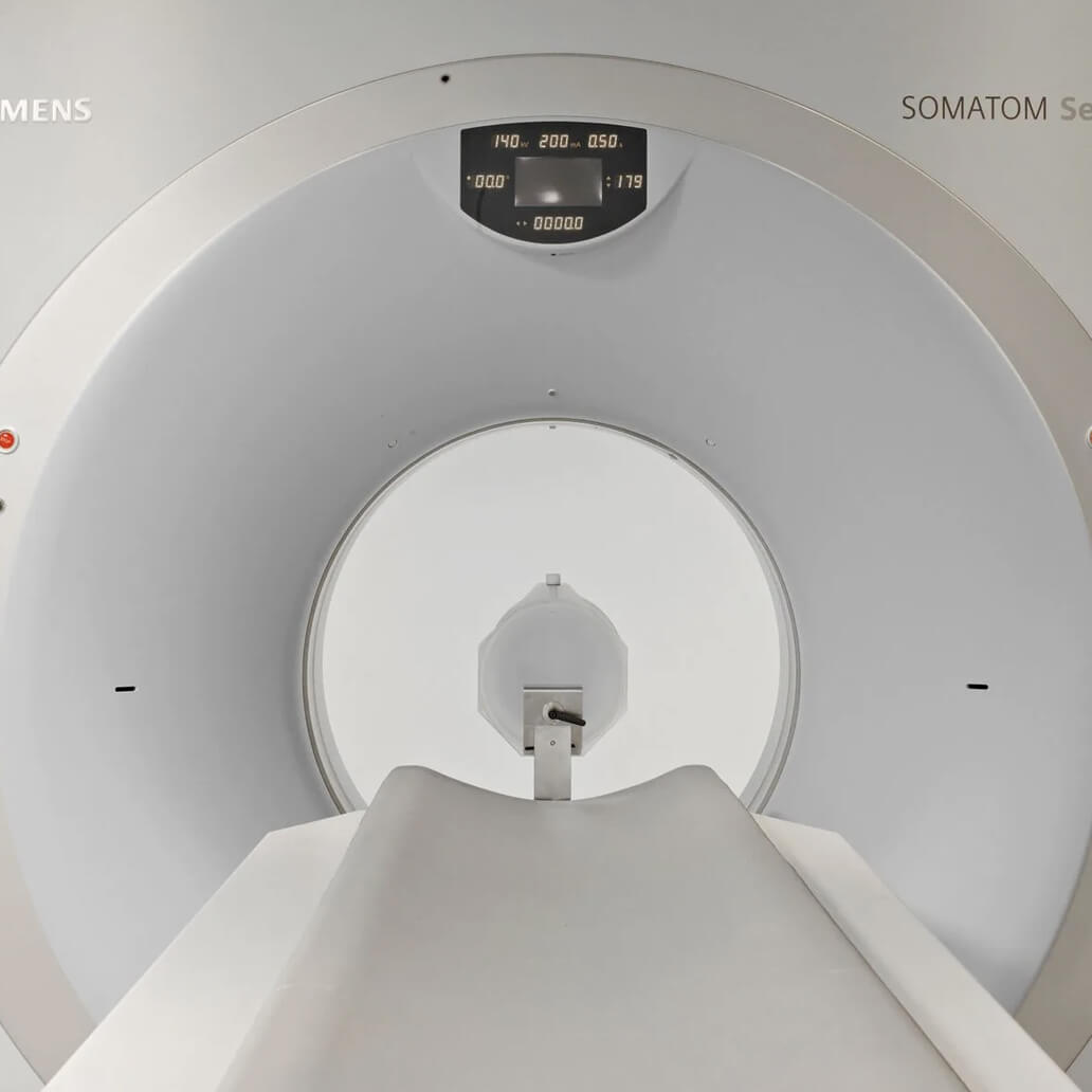 CT scanner tunnel, unoccupied, with a cushioned table leading into the cylindrical opening. Control panel displays numbers. Text reads "SIEMENS" and "SOMATOM Se…". The setting is a medical imaging room.