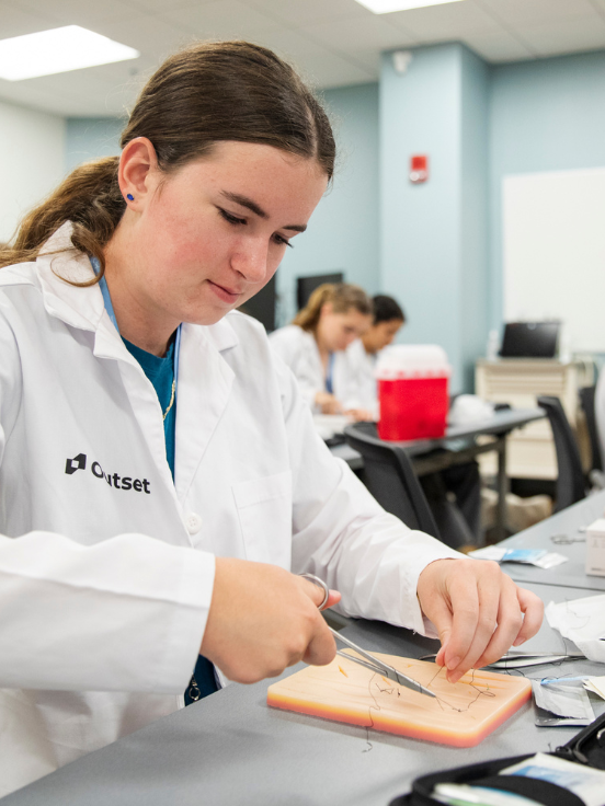 A student interested in medicine in a lab coat practices suturing on a simulation pad at a table, with the "Outset" logo visible, in a classroom with others in the background.