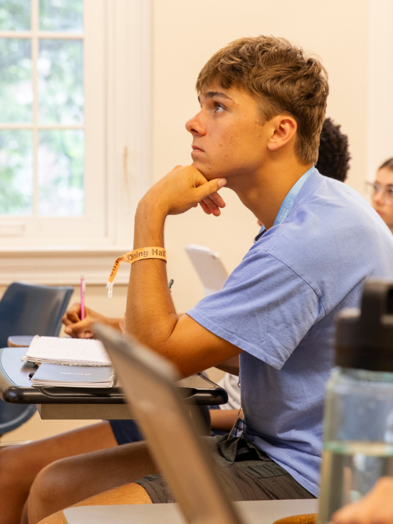 A student attentively listens and takes notes in a classroom. He is wearing a blue shirt and a wristband, sitting at a desk with notebooks. A window is visible in the background.