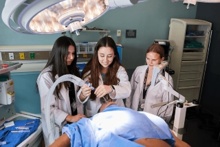 Future medical students in white coats examine a mock patient under bright surgical lights in a hospital operating room. Medical equipment and supplies are visible in the background.